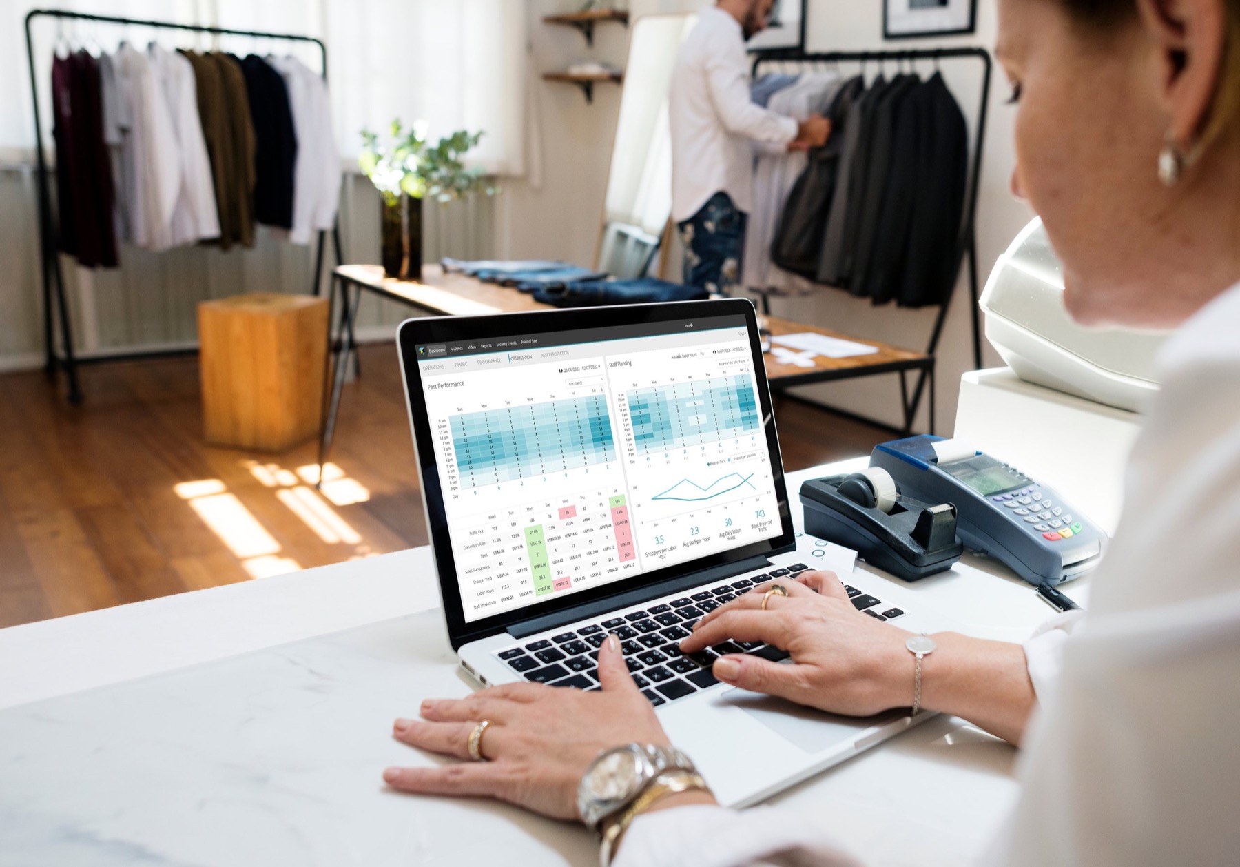 A store associate at a counter working on a laptop that shows the RetailNext staff planning heatmaps, with clothing racks behind her